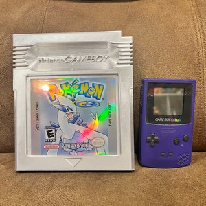 Puede incluir: Un cartucho de juego Game Boy Color blanco con una imagen holográfica de un personaje Pokémon, el texto "Pokémon Versión Plata" y el logotipo de Nintendo. El cartucho está en una caja de plástico transparente. Una consola portátil Game Boy Color morada está al lado del cartucho.