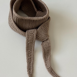 May include: A taupe-colored knitted belt or sash featuring a consistent garter stitch texture throughout. The accessory is coiled into a circular shape with two long, tapered ends hanging down, showcasing a clean, finished edge construction.