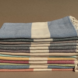 Puede incluir: Una pila de coloridas toallas turcas. Cada toalla presenta un patrón de diamantes y una franja central en un color contrastante. Las toallas son de varios colores, incluyendo azul, verde, amarillo y rojo, con bordes con flecos.