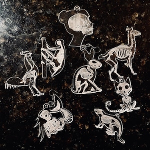 May include: A collection of clear acrylic skeleton-themed charms. The charms include a human head, a rooster, a monkey, a rabbit, a llama, a cat, a mermaid, and a weasel, all depicting skeletal structures. Each charm has a small hole for hanging.
