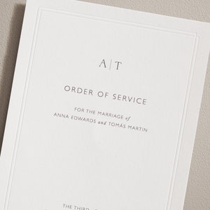 Puede incluir: Un programa de boda blanco con el texto "AT" en una fuente simple en la parte superior. El texto "ORDER OF SERVICE" está centrado debajo de las iniciales. El texto "FOR THE MARRIAGE of ANNA EDWARDS and TOMÁS MARTIN" está centrado debajo del título.