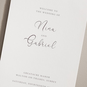 May include: A wedding program with the names "Nina" and "Gabriel" in elegant script. The text "WELCOME TO THE WEDDING OF" is at the top, with the venue and date at the bottom. The program is printed on a light-colored paper.