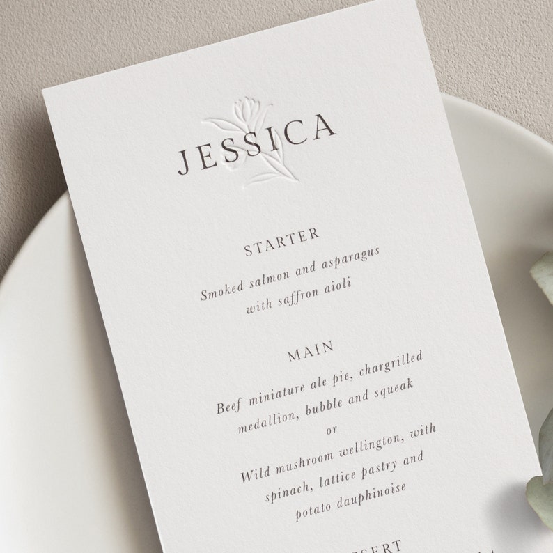 May include: A white wedding menu card with the name "JESSICA" at the top. The menu includes a starter of smoked salmon and asparagus with saffron aioli, and a main course choice of beef miniature ale pie or wild mushroom wellington.