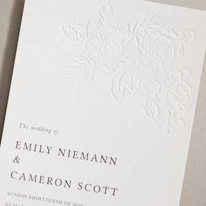 May include: A wedding invitation with a white floral design and the text "The wedding of Emily Niemann & Cameron Scott" in black font. The date "Sunday Eighteenth of May" is also included in black font.