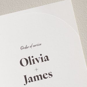 Puede incluir: Una invitación de boda blanca con el texto "Order of service" en una fuente cursiva. Los nombres "Olivia + James" están impresos en negrita, con una fuente sans-serif. La invitación tiene un borde curvo.