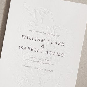 Puede incluir: Invitación de boda con un diseño floral en relieve. El texto dice "Bienvenido a la boda de William Clark & Isabelle Adams, Dieciséis de mayo, Dos mil veintiséis, Iglesia de Santa María, Greatacre."
