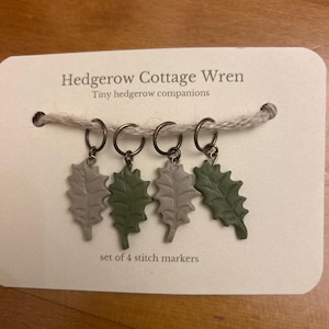 May include: A set of 4 stitch markers on a cream-coloured card labelled Hedgerow Cottage Wren. Each marker features a bronze-toned ring attached to a leaf-shaped charm in alternating shades of sage green and muted grey, with textured vein details.