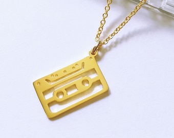 May include: A gold-toned necklace featuring a pendant shaped like a cassette tape. The pendant is a flat, cutout design with two circular holes and a rectangular center. The necklace chain is a delicate gold-toned link style.
