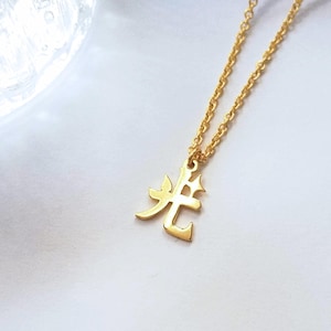 May include: A gold-colored necklace featuring a delicate chain and a pendant shaped like the Japanese kanji character for light, which is 'Hikari'. The pendant has a polished finish and is suspended from a thin, matching gold-colored link chain.