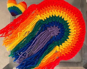 May include: A rainbow crocheted wall hanging with a fringe of yarn in red, orange, yellow, green, blue, indigo, and violet. A small crocheted heart with a rainbow pattern is attached to the top of the hanging.