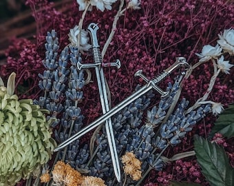 May include: Two silver swords with intricate details are crossed against a backdrop of purple and lavender flowers. The swords have a sharp point and a curved handle.