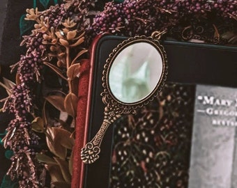 May include: A vintage-style hand mirror with an ornate metal frame and a clear glass surface. The mirror is resting on a book with a floral design.