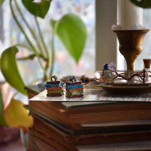 Puede incluir: Dos pilas en miniatura de libros coloridos, cada una con una pequeña figura en la parte superior, se muestran en una pila de libros antiguos. Los libros están sobre una mesa con un candelero y un plato de vidrio con piedras de colores.