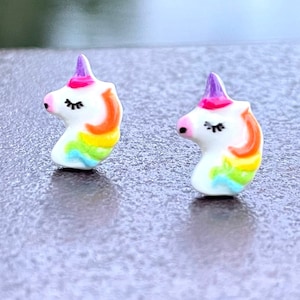 May include: A pair of small unicorn earrings with a rainbow mane and a purple horn. The unicorn's eyes are closed and the earrings are made of white ceramic.
