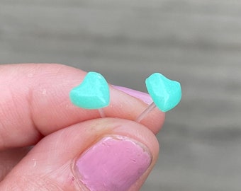 Op de afbeelding: Een paar kleine, mintgroene hartvormige oorstekers. De oorbellen worden tussen de vingers gehouden, wat hun delicate ontwerp laat zien. De oorbellen zijn gemaakt van een doorschijnend materiaal.