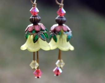 Op de afbeelding: Een paar gele, bloemvormige oorbellen met groene bladeren, roze accenten en kristallen druppels.