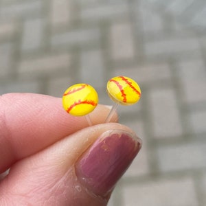 May include: A pair of small, yellow and red softball earrings. The earrings are on a clear post.