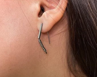 Può includere: Primo piano di un orecchio con un orecchino di perline d'argento. L'orecchino è un pezzo lungo e sottile di metallo con piccole perline attaccate. L'orecchino è appeso al lobo dell'orecchio.