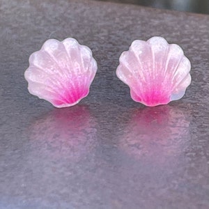 May include: A pair of pink and white seashell shaped earrings. The earrings are made of resin and have a glossy finish.