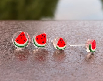 Könnte beinhalten: Drei Paar Wassermelonen-Scheiben-Ohrstecker. Die Ohrringe sind aus klarem Kunststoff mit einem roten Wassermelonen-Scheiben-Design.