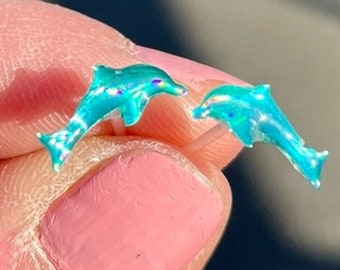 Op de afbeelding: Een paar kleine, turquoise dolfijn oorbellen. De oorbellen worden tussen de vingers gehouden, waarbij hun iriserende afwerking wordt getoond. De dolfijnen zijn gedetailleerd met vinnen en ogen, die licht reflecteren.