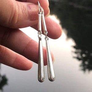 May include: Silver teardrop earrings with a chain detail. The earrings are hanging from a person's hand.