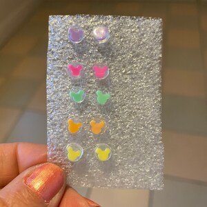 May include: A set of colorful stud earrings in a variety of colors, including purple, pink, green, orange, and yellow. Each earring features a small, round design with a Mickey Mouse head shape. The earrings are displayed on a clear, textured backing.