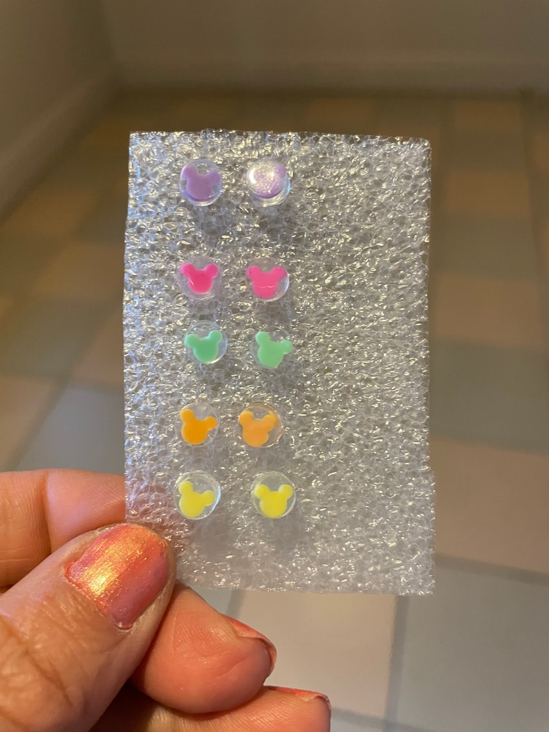 May include: A set of colorful stud earrings in a variety of colors, including purple, pink, green, orange, and yellow. Each earring features a small, round design with a Mickey Mouse head shape. The earrings are displayed on a clear, textured backing.
