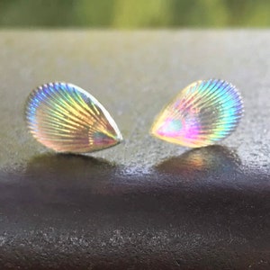 May include: Two iridescent, teardrop-shaped earrings with a scalloped design. The earrings are a light, pearly white with rainbow-like reflections.