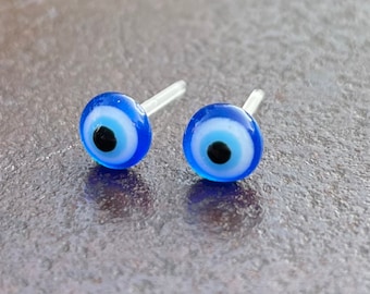 Op de afbeelding: Een paar kleine blauwe boze oog oorbellen met witte en zwarte accenten. De oorbellen liggen op een grijs oppervlak.