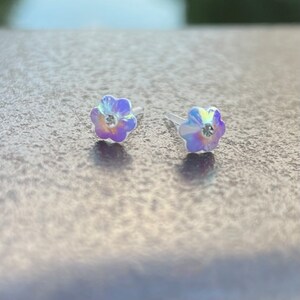Pu&ograve; includere: Un paio di orecchini a bottone a forma di fiore iridescente con un cristallo trasparente al centro di ogni fiore.