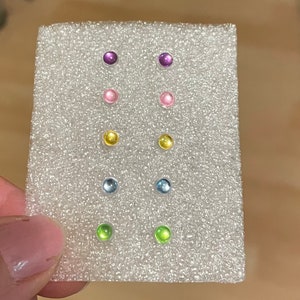 May include: A white foam board with 10 small round earrings in various colors. The earrings are arranged in pairs, with each pair in a different color. The colors are purple, pink, yellow, blue, and green.