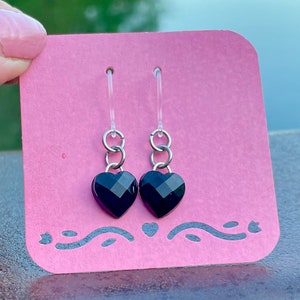 May include: A pair of black heart-shaped earrings with silver hoops and chains. The earrings are displayed on a pink card with a decorative border.
