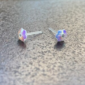 Pu&ograve; includere: Un paio di orecchini a bottone a forma di fiore iridescente con strass trasparenti al centro. Gli orecchini sono su uno sfondo bianco.