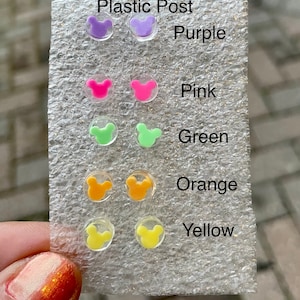 May include: A selection of hypoallergenic plastic post earrings in various colors, including purple, pink, green, orange, and yellow. Each earring features a small, cartoon mouse head design. The colors are labeled on the packaging.