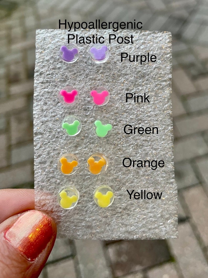 May include: A selection of hypoallergenic plastic post earrings in various colors, including purple, pink, green, orange, and yellow. Each earring features a small, cartoon mouse head design. The colors are labeled on the packaging.