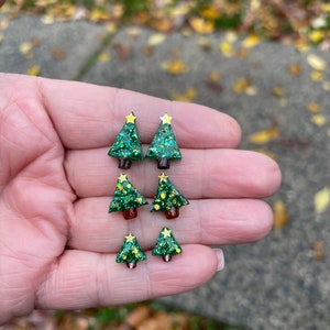 May include: A set of four green glitter Christmas tree earrings with gold star accents. The earrings are different sizes and are displayed on a hand.