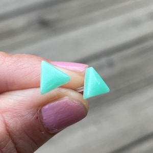 May include: A pair of small, light green triangle shaped earrings. The earrings are made of a smooth, opaque material.