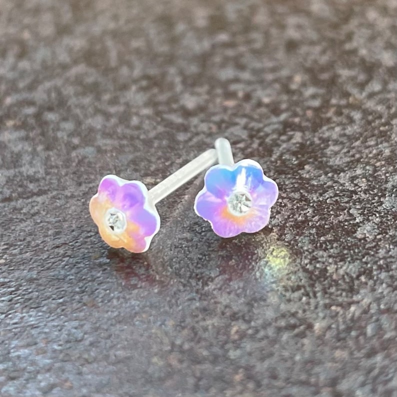 Pu&ograve; includere: Un paio di orecchini a perno a forma di fiore iridescente con strass trasparenti al centro. Gli orecchini sono su una superficie grigia testurizzata.