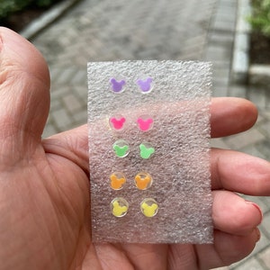 May include: A set of six pairs of colorful stud earrings in a clear plastic package. Each pair features a Mickey Mouse head design in various colors, including purple, pink, green, orange, and yellow. The earrings are small and appear to be made of acrylic.