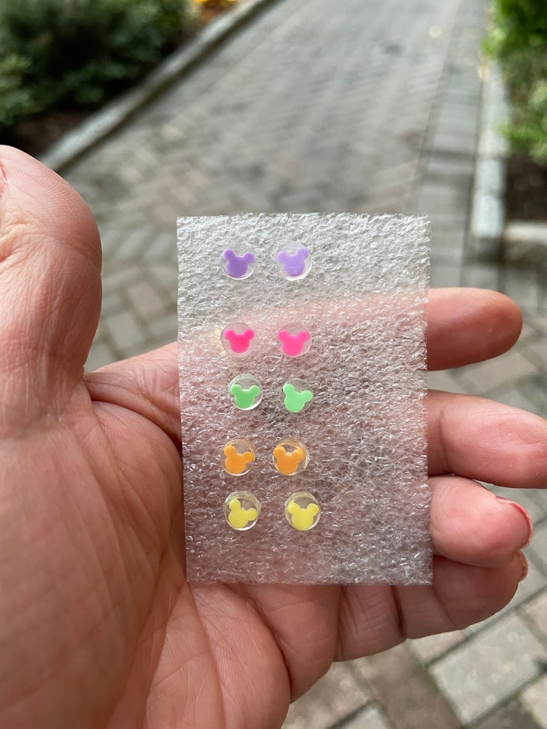 May include: A set of six pairs of colorful stud earrings in a clear plastic package. Each pair features a Mickey Mouse head design in various colors, including purple, pink, green, orange, and yellow. The earrings are small and appear to be made of acrylic.