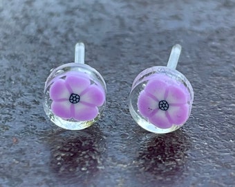 Op de afbeelding: Een paar kleine, heldere glazen oorbellen met een paars bloemenmotief. De bloemen hebben een wit centrum en een zwarte omtrek.