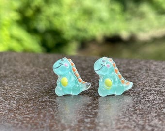 Op de afbeelding: Een paar turquoise blauwe dinosaurus oorbellen met oranje stippen en gele ogen. De dinosaurussen kijken naar voren met hun mondjes lichtjes open.