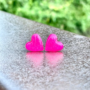 May include: A pair of pink heart-shaped stud earrings. The earrings are made of a shiny, sparkly material.