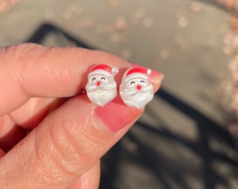 Op de afbeelding: Een paar witte kerstman oorbellen met rode hoeden en glitter accenten.