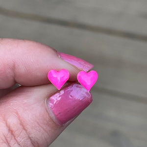 Puede incluir: Un par de pendientes de botón en forma de corazón rosa. Los pendientes son pequeños y delicados, y son perfectos para añadir un toque de fantasía a cualquier atuendo.