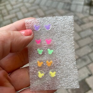 May include: A collection of six pairs of colorful, clear acrylic stud earrings. Each pair features a Mickey Mouse head shape in shades of purple, pink, green, orange, and yellow. The earrings are displayed on a clear plastic backing.