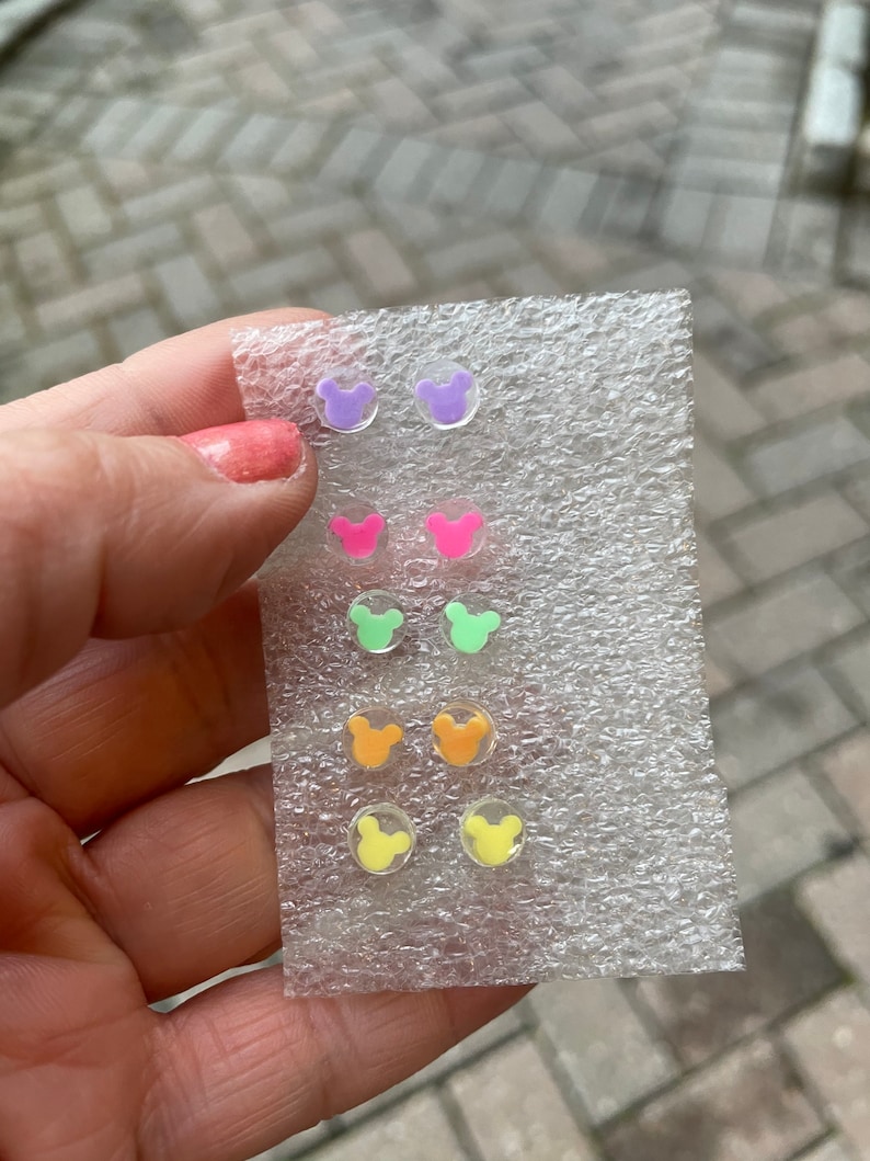 May include: A collection of six pairs of colorful, clear acrylic stud earrings. Each pair features a Mickey Mouse head shape in shades of purple, pink, green, orange, and yellow. The earrings are displayed on a clear plastic backing.