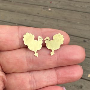 May include: Two gold-colored metal chicken charms. The charms are in the shape of a chicken with a ruffled tail and a small hole at the top for hanging.