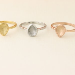 May include: Three teardrop-shaped rings with a bezel setting. The rings are gold, silver, and rose gold.